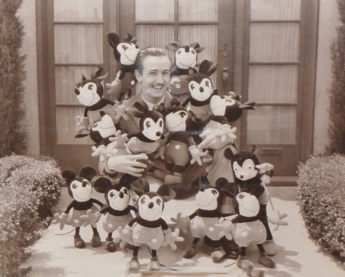 Image shows a black and white photograph of Walt Disney surrounded by many Mickey Mouse plush toys.