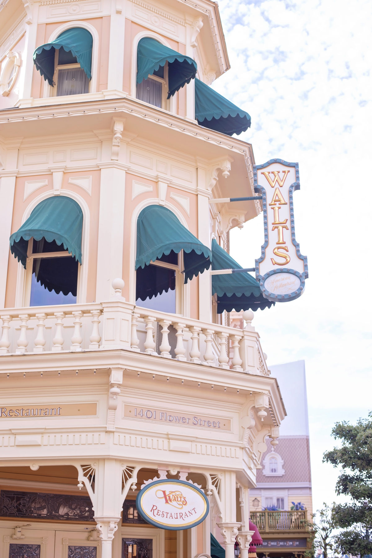 Image shows the exterior of Walt's Restaurant on Main Street, Disneyland Paris in the daytime. The building is pink and the sign 1402 flower street can be seen above the sign.  Green fabric awning shutters are over the windows.