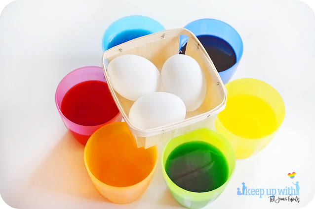 Image shows equipment needed to dye eggs for easter.  White eggs in a wooden berry basket and six cups of food coloured water - pink/red, orange, yellow, green blue and purple.