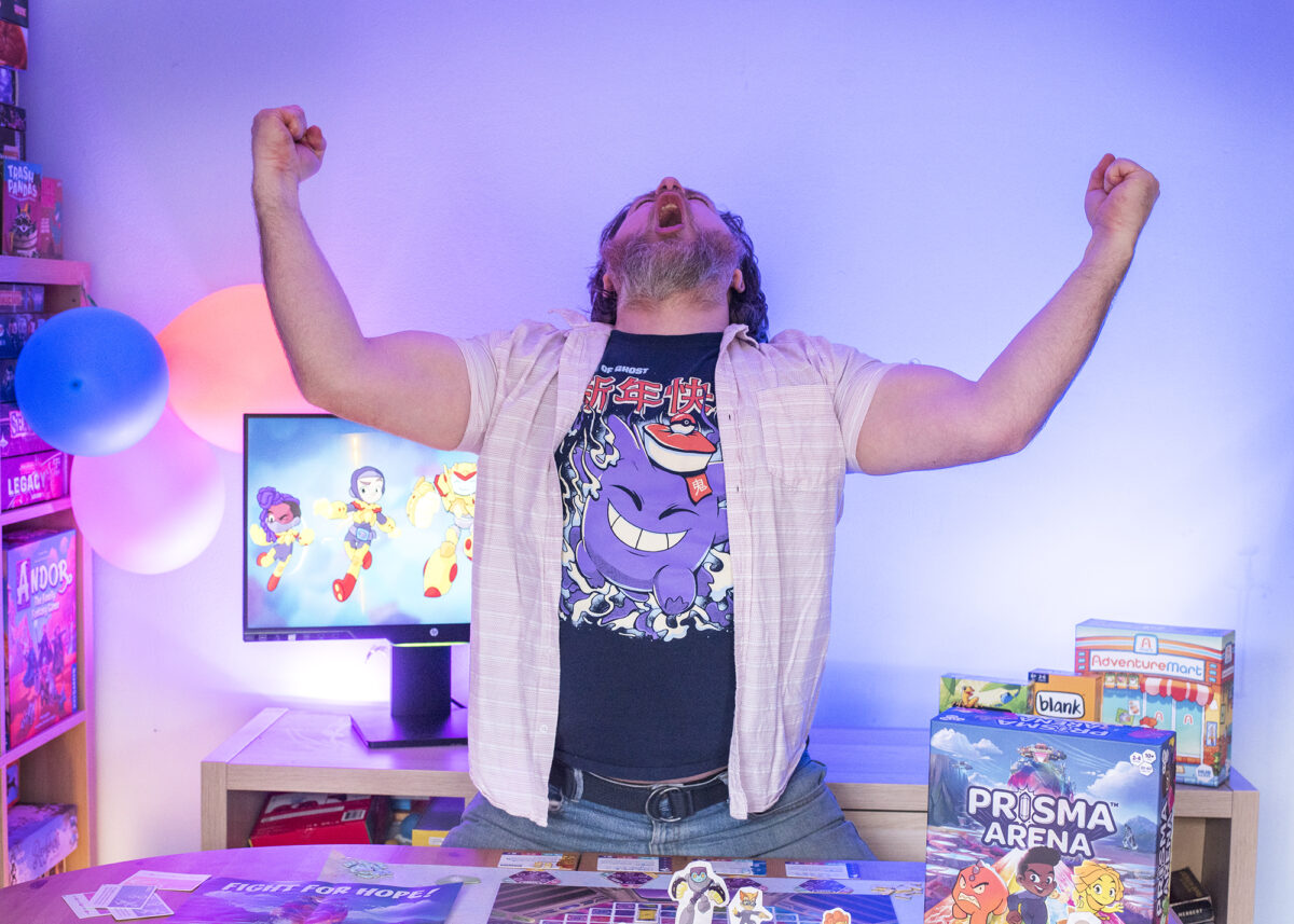 Photo shows man with arms in air triumphantly over a copy of Prisma Arena from Hub Games on the table in front of him.  The lighting is blue and purple.