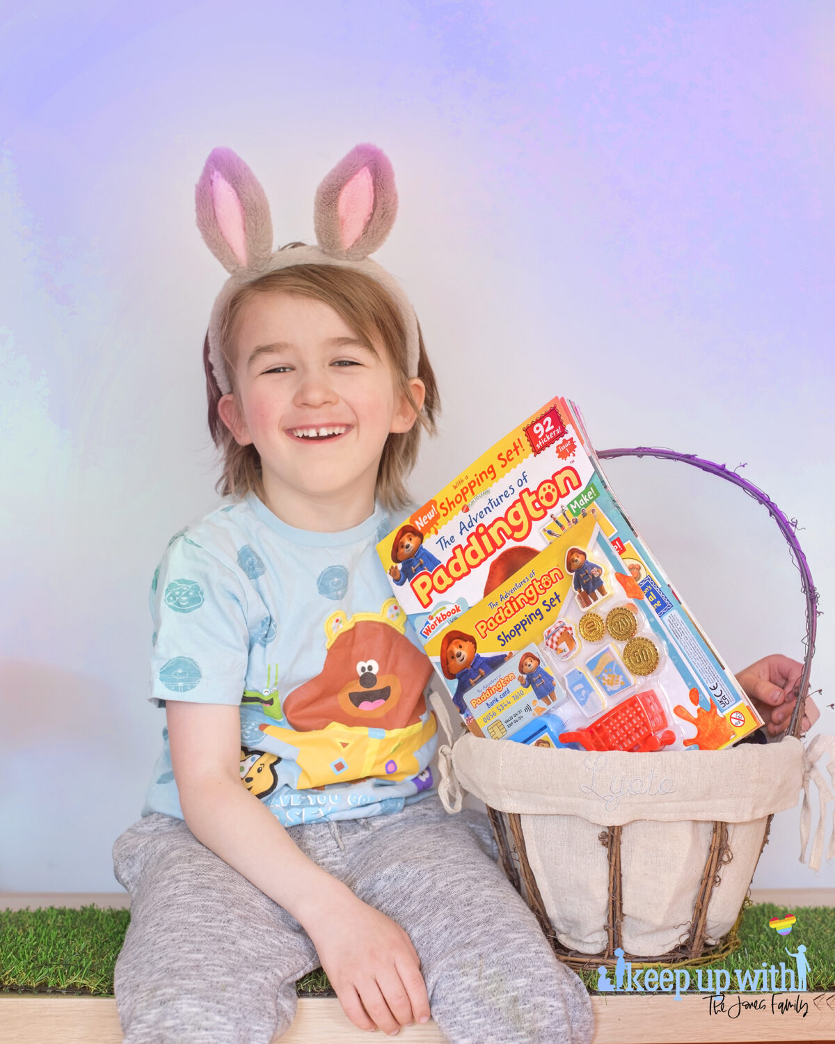 The Adventures of Paddington Bear Magazine EYFS. Photograph shows a boy with grey and pink rabbit ears wearing grey tacksuit bottoms and an aqua coloured Hey Duggee Cbeebies t-shirt for children in need 2020.  The boy is sat on artificial grass and holding an easter basket which contain the new Adventures of paddington magazine. He is smiling.  