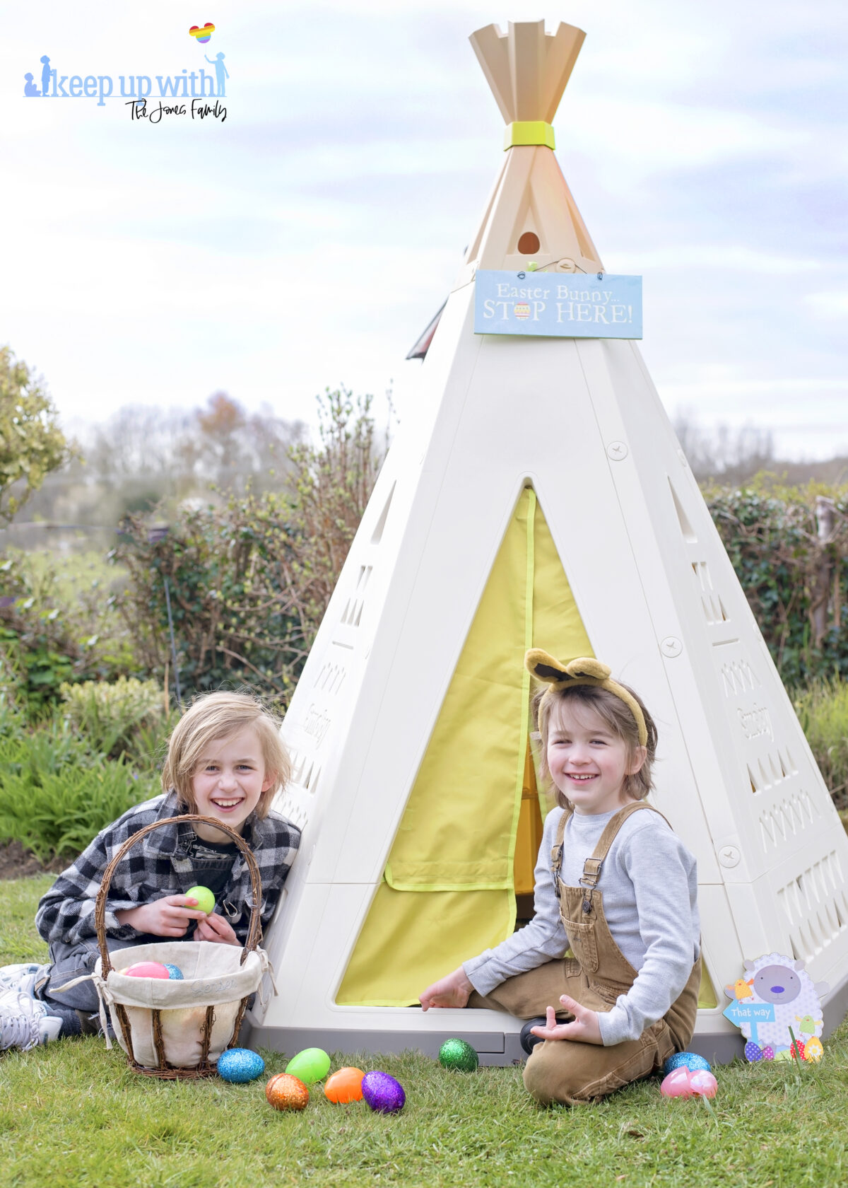 Image shows the new Smoby Teepee, available on Amazon, in the garden. The teepee is cream, white and green with a triangle pattern on the sides.  There is a fabric door which is lime green and two little boys are sat on the outside of the teepee, dressed for easter with bunny ears on.  They are laughing.  They have easter baskets filled with coloured easter eggs which have spilled onto the floor.  The teepee is decorated with a sign which reads Easter Bunny Stop here!
