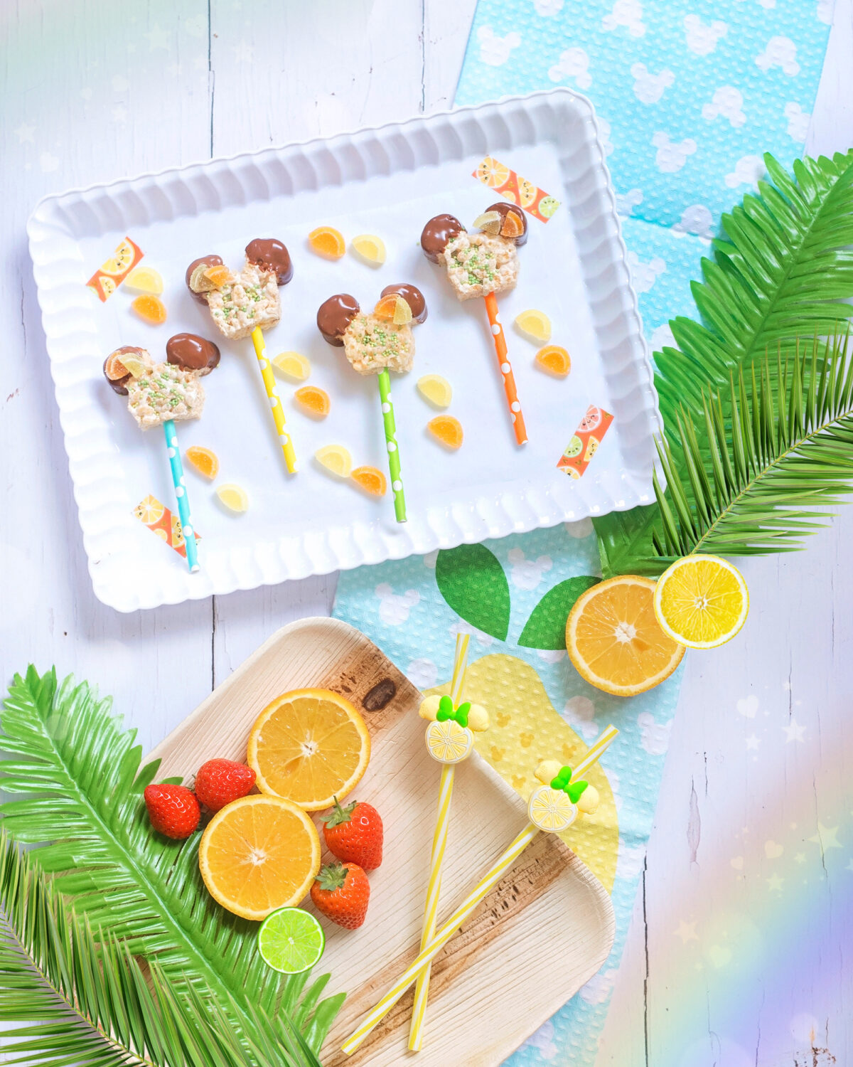 Image shows white wooden backdrop with a white tray of rice krispie treats in the shape of mickey mouse's head on coloured straws.  The Mickey Treats are dipped in chocolate and covered in sprinkles. There are orange and lemon jelly pieces on the ears. Around the tray are orange, lime and lemon slices, tropical leaves and a rainbow overlay.  This is Disney fun food.  Image by Keep Up With The Jones Family.