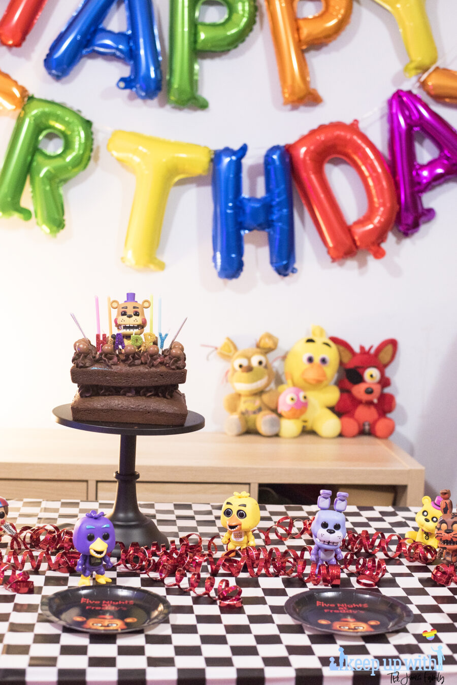 How to throw a Five Nights at Freddy's Party. Image shows a party table covered in a black checkered tablecloth, happy birthday spelled out in rainbow coloured balloons as a garland across the wall, three FNAF plushies; Foxy, Chica and Plushtrap sat on the sideboard and a chocolate cake on a black cake pedestal with a Funko Pop Viny; Rockstar Freddy on top of it.