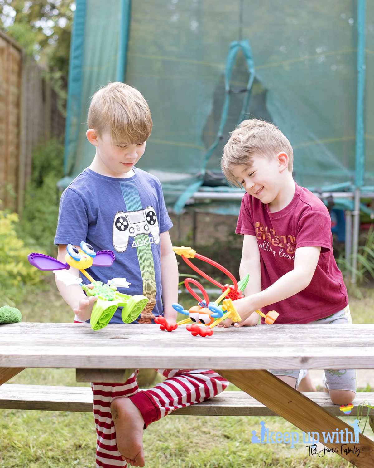 Image shows two boys playing with toys whilst sat at a wooden picnic table in the garden.  On the wooden picnic table in front of them are two creatures which are made from pieces of the Kid K'nex Budding Builders construction toys box from Basic Fun UK.  By Keep Up With The Jones Family.