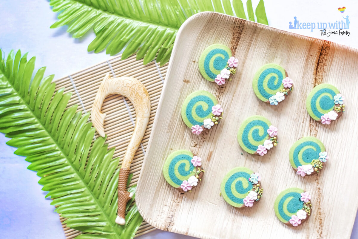 Image shows how to make Disney's Moana Heart of Te Fiti Biscuits, a swirl of bright and dark green, dipped slightly in milk chocolate and embellished with sugar blossom flowers and green sprinkles.  They are set on a bamboo plate with a fern underneath, and a small toy version of Maui's fish hook is resting next to the plate also. Image by keep up with the jones family.