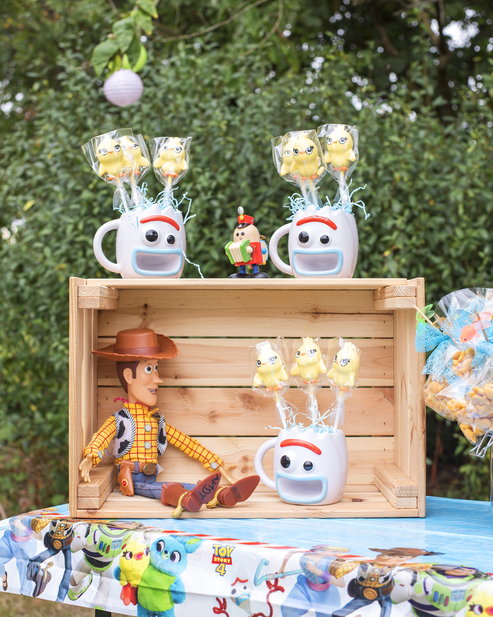 How to host a toy story party: Image shows a party table with a toy story tablecloth and wooden crate perched on top. Woody the Disney Cowboy toy sits inside the crate and three mugs in the shape of the face of Forky from Toy Story Four are sat on and in the crate. Inside the mugs are Ducky cake pops. The table is outdoors at a toy story party.