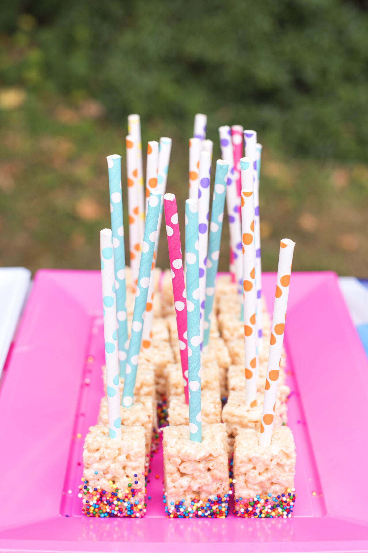 Image shows a bright pink plastic party platter filled with Rice Krispie treat lollipops dipped in rainbow sprinkles. The lollipops have coloured paper straw handles.