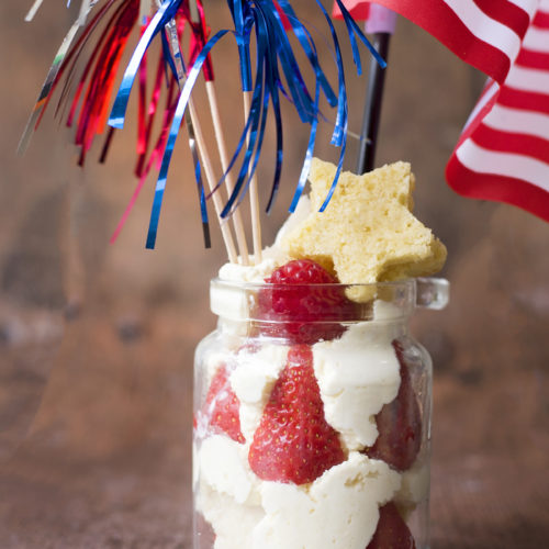 Image shows a 4th July Strawberry Cake Jar dessert on a dark wooden tabletop. The jar contains piped whipped cream and strawberries, with stars cut out of vanilla angel cake inside and on top. There is an american flag and three metallic firework sparklers in red, white and blue sticking out of the jar. Image by Sara-Jayne Jones of Keep Up With The Jones Family.