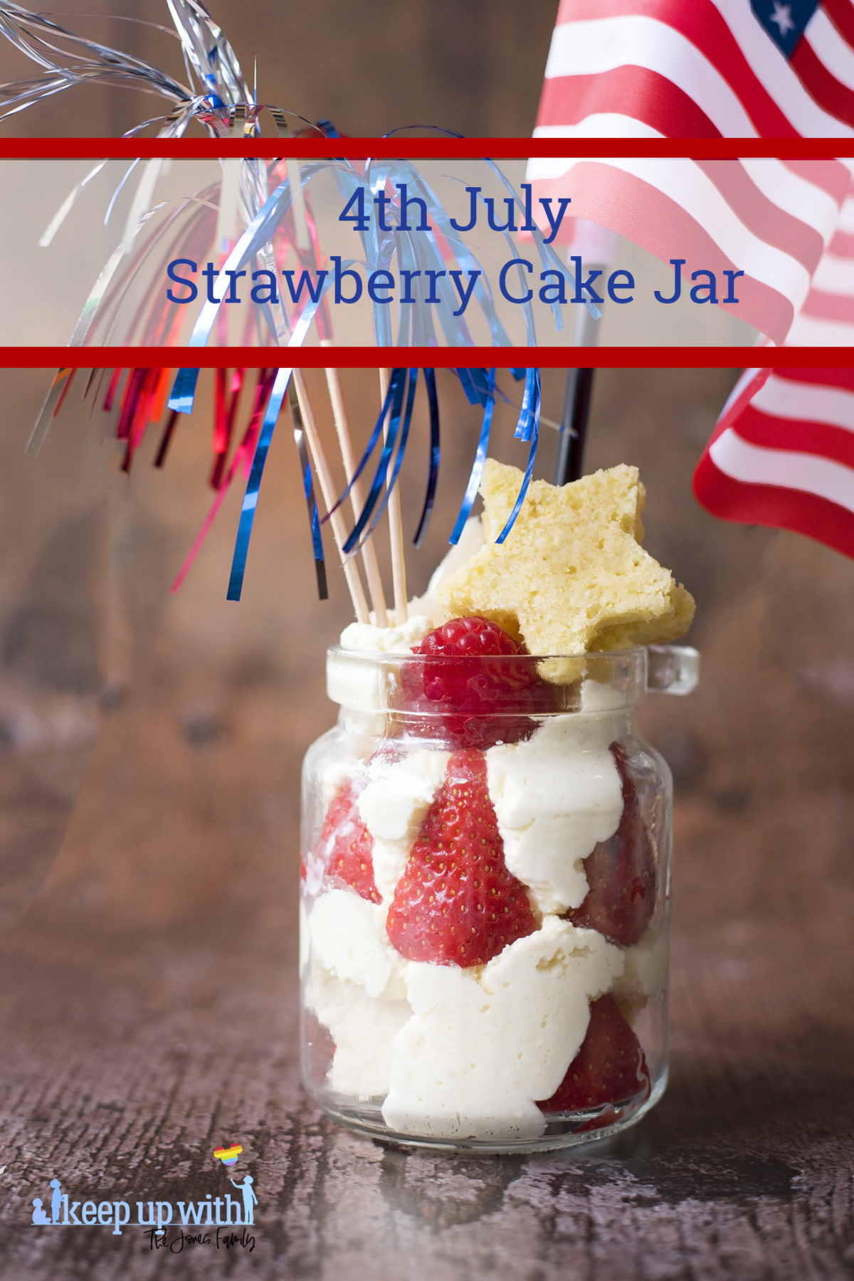 Image shows a 4th July Strawberry Cake Jar dessert on a dark wooden tabletop. The jar contains piped whipped cream and strawberries, with stars cut out of vanilla angel cake inside and on top. There is an american flag and three metallic firework sparklers in red, white and blue sticking out of the jar. Image by Sara-Jayne Jones of Keep Up With The Jones Family.