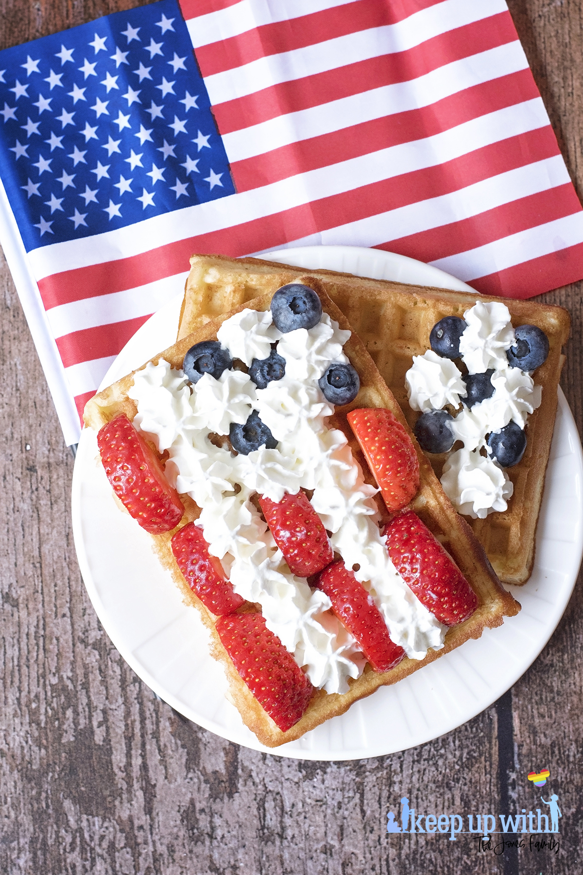 Image shows rectangular sweet breakfast waffles on a white vera wang plate. The waffles are being decorated in the style of the American Flag. There are sliced strawberries for the red stripes, blueberries for the space around the stars, and cream is piped in star shapes as the white stars and stripes. The plate is set on a dark hardwood background and there is a small American Flag next to it. Image by Sara-Jayne Jones, Keep Up With The Jones Family.