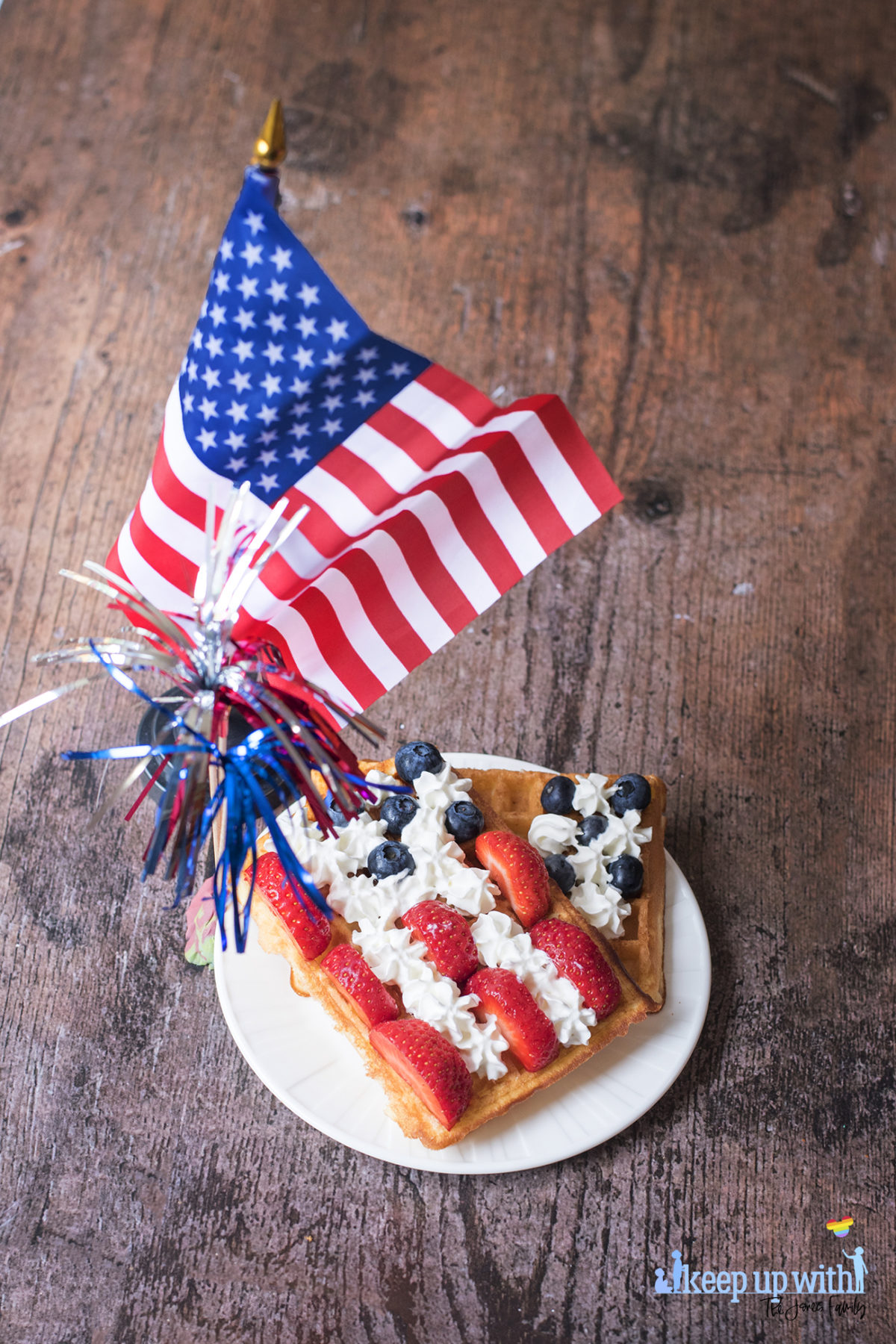 Image shows rectangular sweet breakfast waffles on a white vera wang plate. The waffles are decorated in the style of the American Flag. There are sliced strawberries for the red stripes, blueberries for the space around the stars, and cream piped as the white stars and stripes. The plate is set on a dark hardwood background and there is a small American Flag next to it and a red, silver and blue set of firework decorations. Image by Sara-Jayne Jones, Keep Up With The Jones Family.
