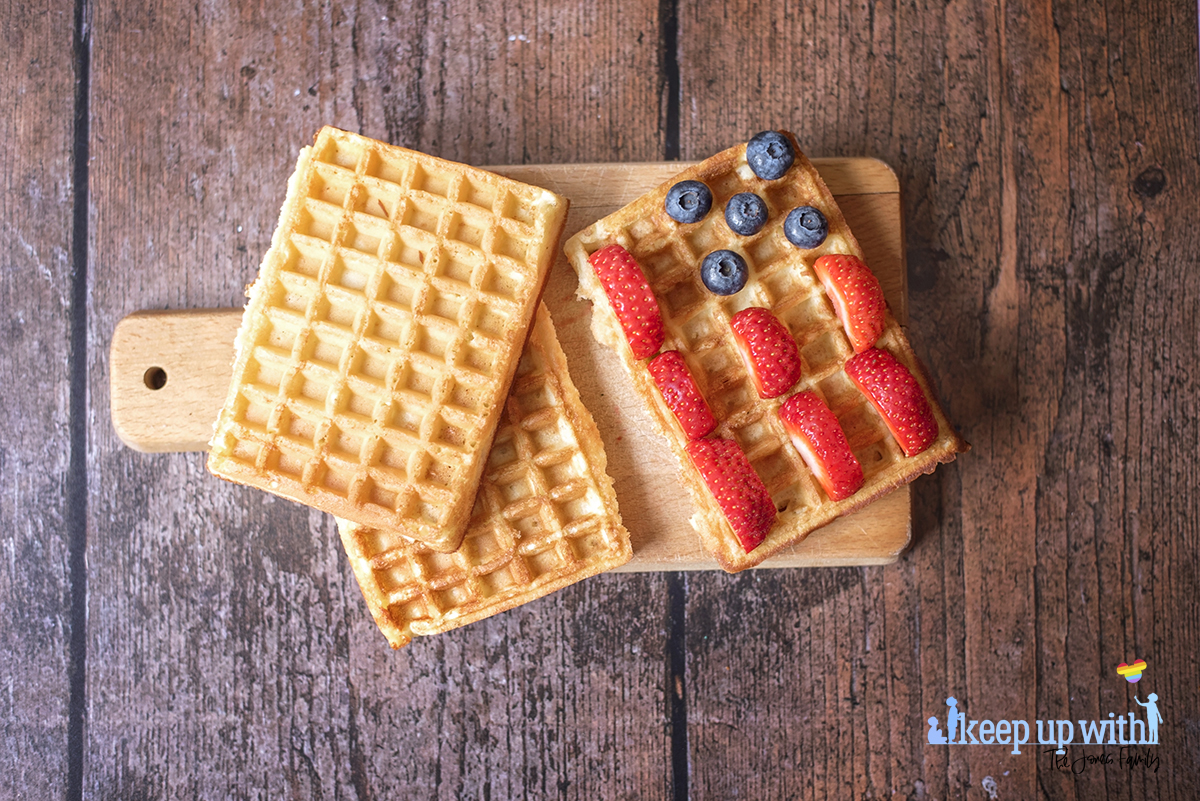 Image shows rectangular sweet breakfast waffles on a small wooden chopping paddle. The waffles are being decorated in the style of the American Flag. There are sliced strawberries for the red stripes, blueberries for the space around the stars, and they are waiting for cream to be piped as the white stars and stripes. The plate is set on a dark hardwood background. Image by Sara-Jayne Jones, Keep Up With The Jones Family.