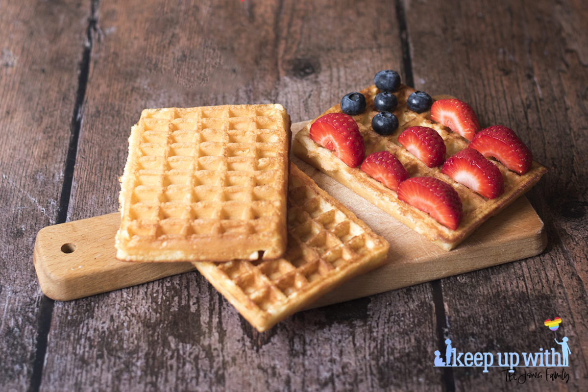 Image shows rectangular sweet breakfast waffles on a small wooden chopping paddle. The waffles are being decorated in the style of the American Flag. There are sliced strawberries for the red stripes, blueberries for the space around the stars, and they are waiting for cream to be piped as the white stars and stripes. The plate is set on a dark hardwood background. Image by Sara-Jayne Jones, Keep Up With The Jones Family.