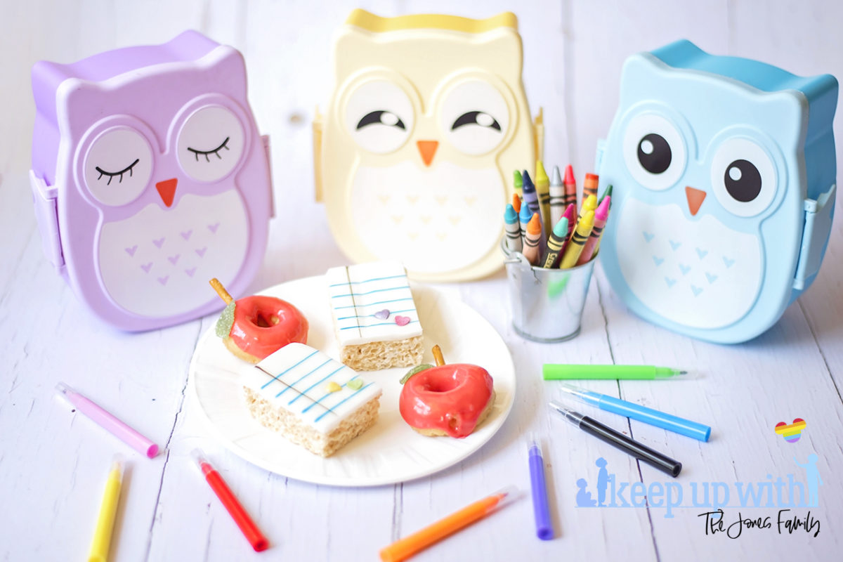 Image shows Back to School Rice Krispie treats and back to school apple shaped doughnuts on a white Vera Wang for Wedgwood plate. There are three pastel coloured owl bento boxes in the background and a small tin bucket of Crayola crayons alongside it. Cake pens surround the plate on the white wooden background. Image by Sara-Jayne from Keep Up With The Jones Family.