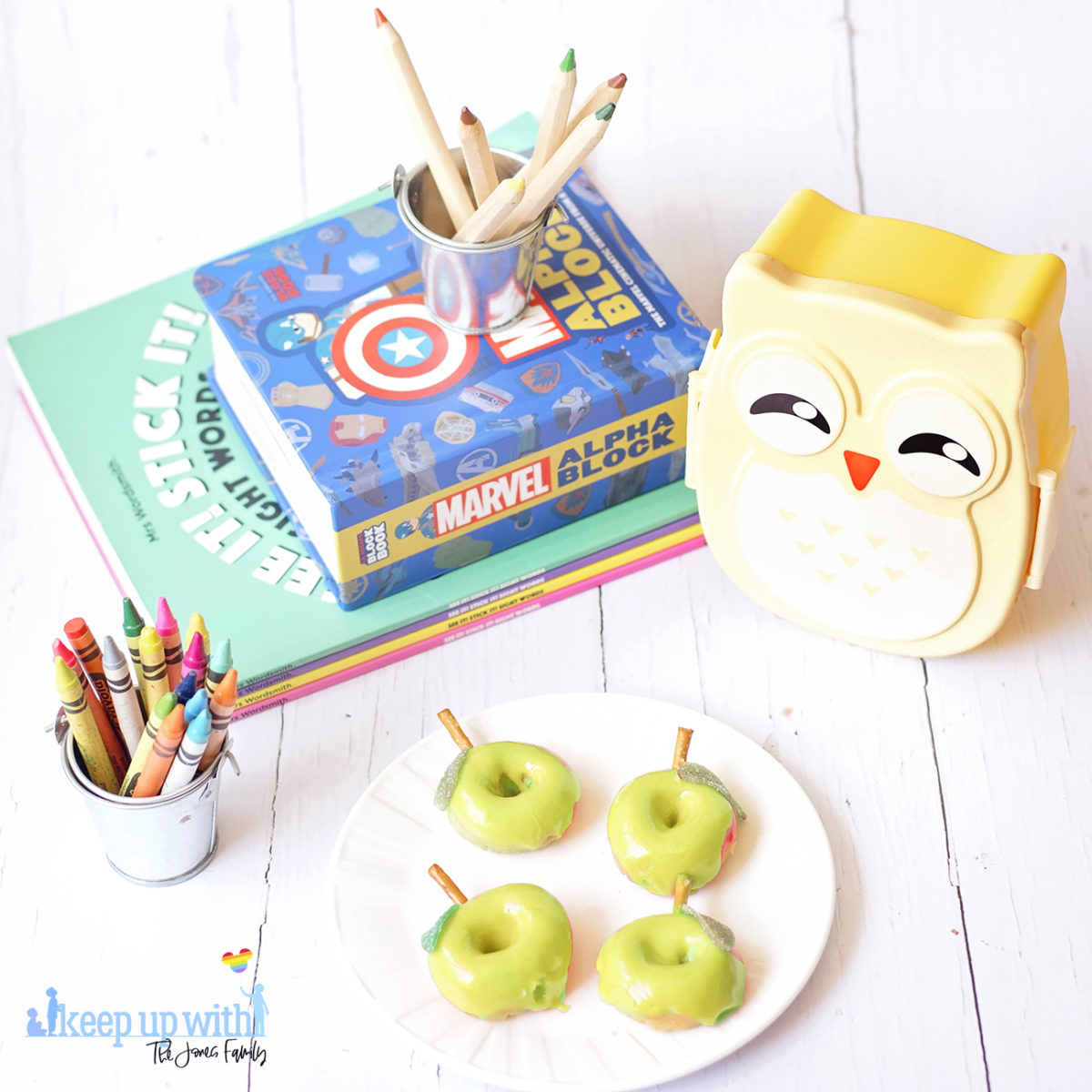 Image shows green apple shaped Back to School Doughnuts on a white Vera Wang plate, sitting on a white wooden surface. There are two small owl shaped bento boxes and books in the background, along with a bucket of crayola crayons. Image by Sara-Jayne from Keep Up With The Jones Family.