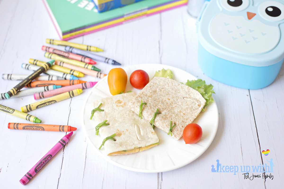 Image shows Book Sandwich Wraps on a white Vera Wang plate, resting on a white wooden surface surrounded by a blue owl lunchbox, a tin bucket of crayola crayons in various colours and a stack of books including Mrs Wordsmith and Marvel Alphablock. Image by Sara-Jayne of Keep Up With the Jones Family.