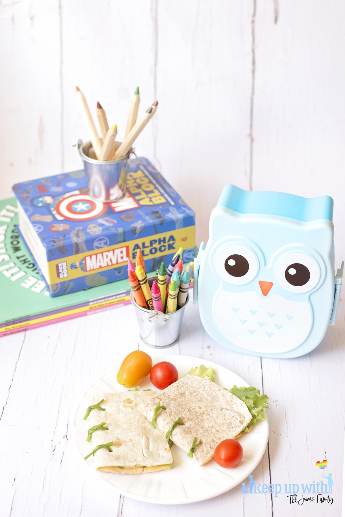 Image shows Book Sandwich Wraps on a white Vera Wang plate, resting on a white wooden surface surrounded by a blue owl lunchbox, a tin bucket of crayola crayons in various colours and a stack of books including Mrs Wordsmith and Marvel Alphablock. Image by Sara-Jayne of Keep Up With the Jones Family.