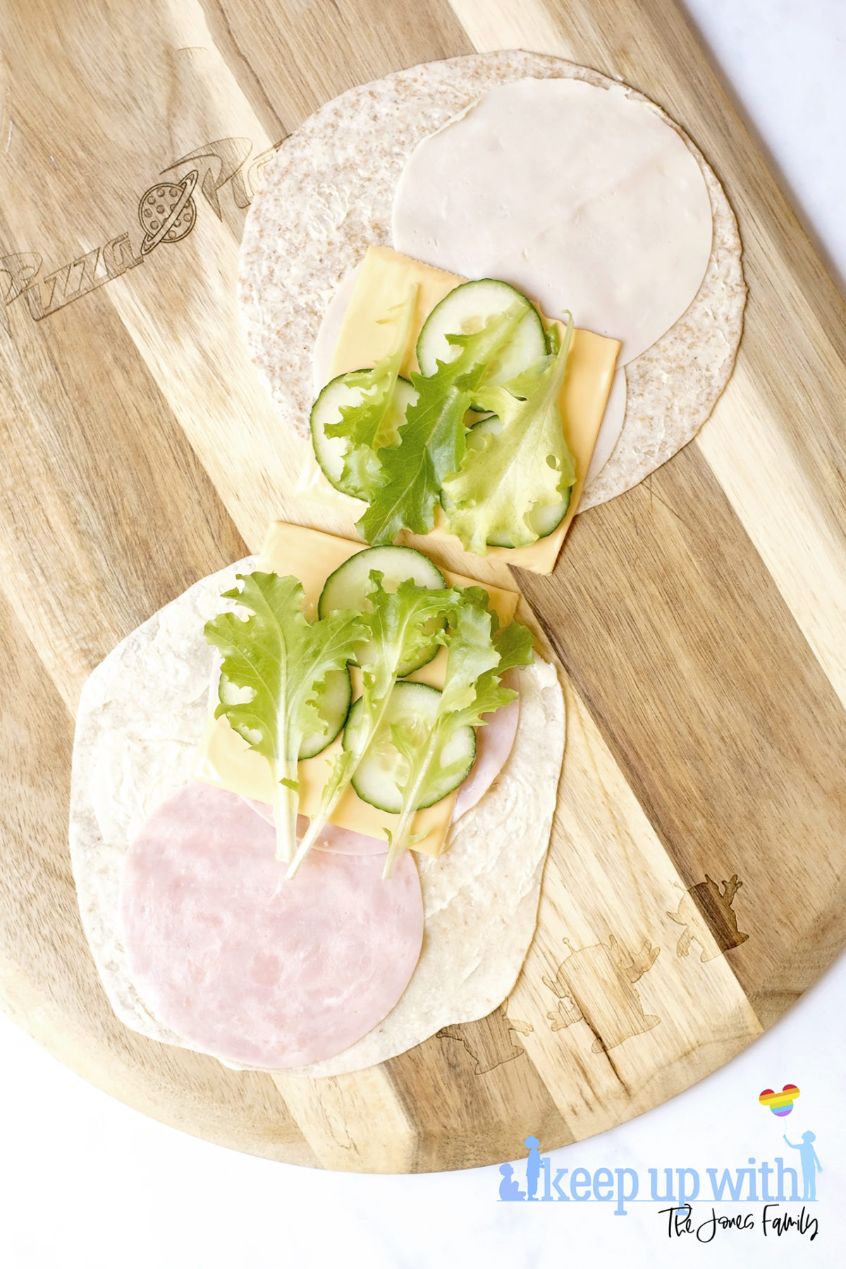 Image Shows step one of creating Book Sandwich Wraps. On a wooden chopping board sit two mini wraps, with ham, chicken, lettuce, cheese slices and cucumber on them. Image by Sara-Jayne of Keep Up With The Jones Family