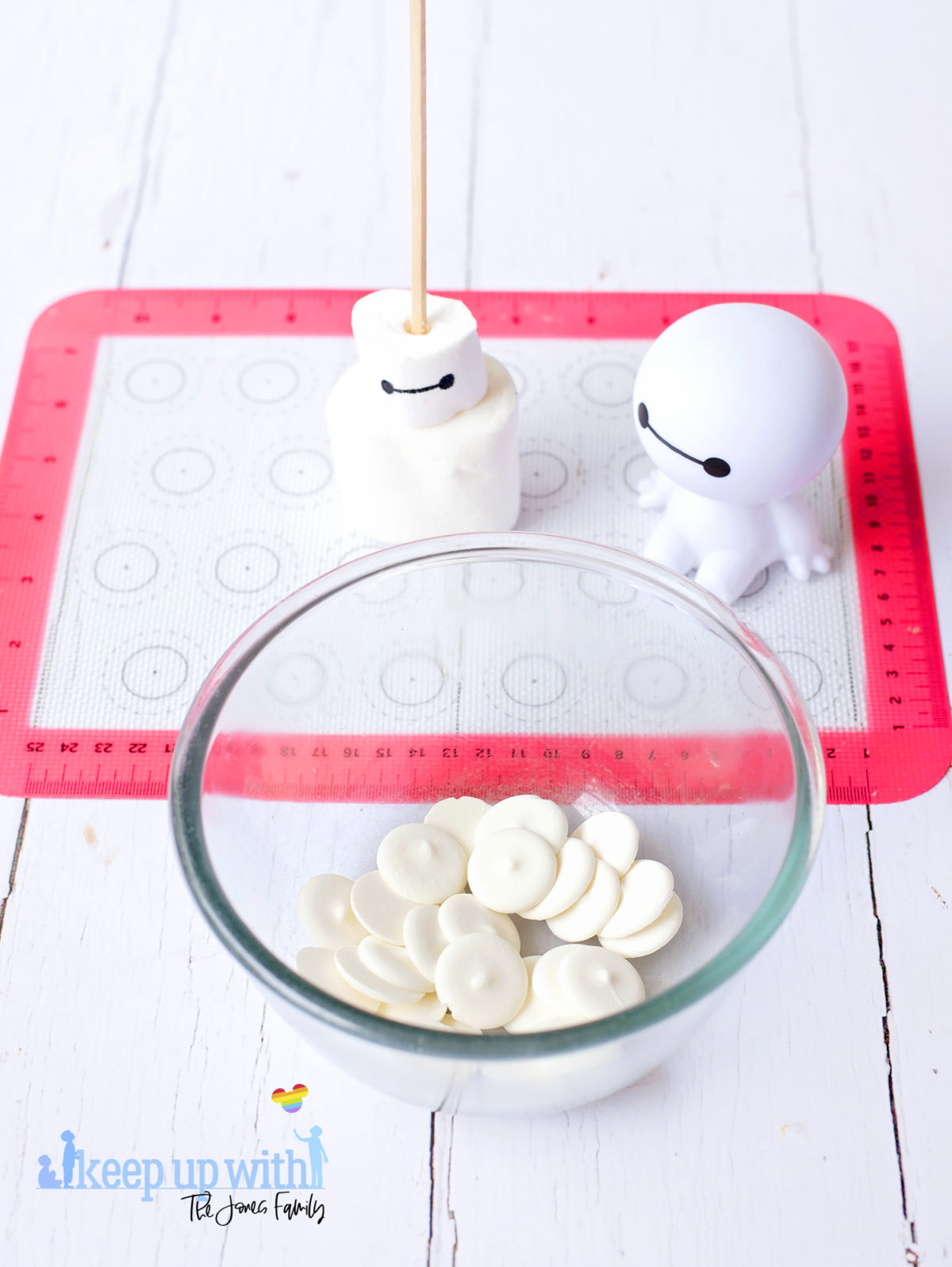 Image shows a plastic Baymax figure from Disney Pixar's Big Hero 6, and a Marshmallow Bayman Pop sat next to it on a red and white silicone baking mat. In front of them is a bowl full of white candy melts in a glass microwaveable bowl. Image by Keep Up With The Jones Family.