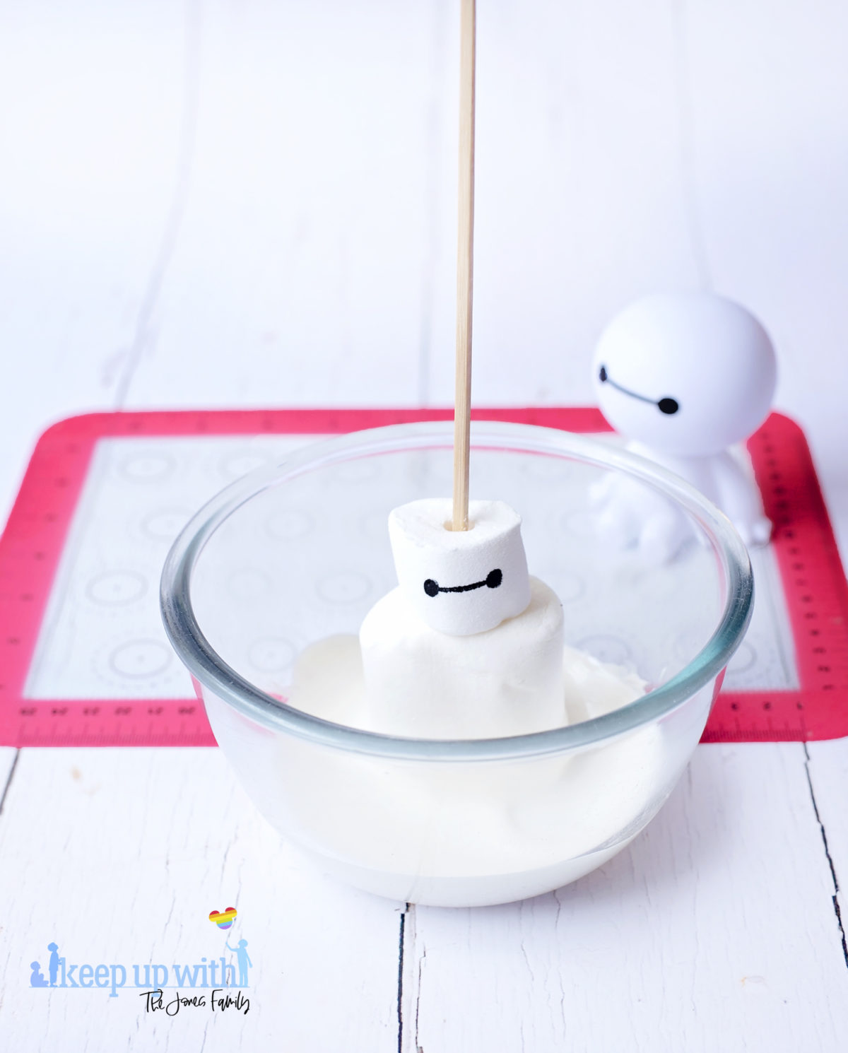 Image shows a plastic Baymax figure from Disney Pixar's Big Hero 6 sat on a white tabletop. In front of him is a glass bowl containing heated white candy melts and a Baymax Marshmallow Pop, being coated. Image by keep up with the jones family.