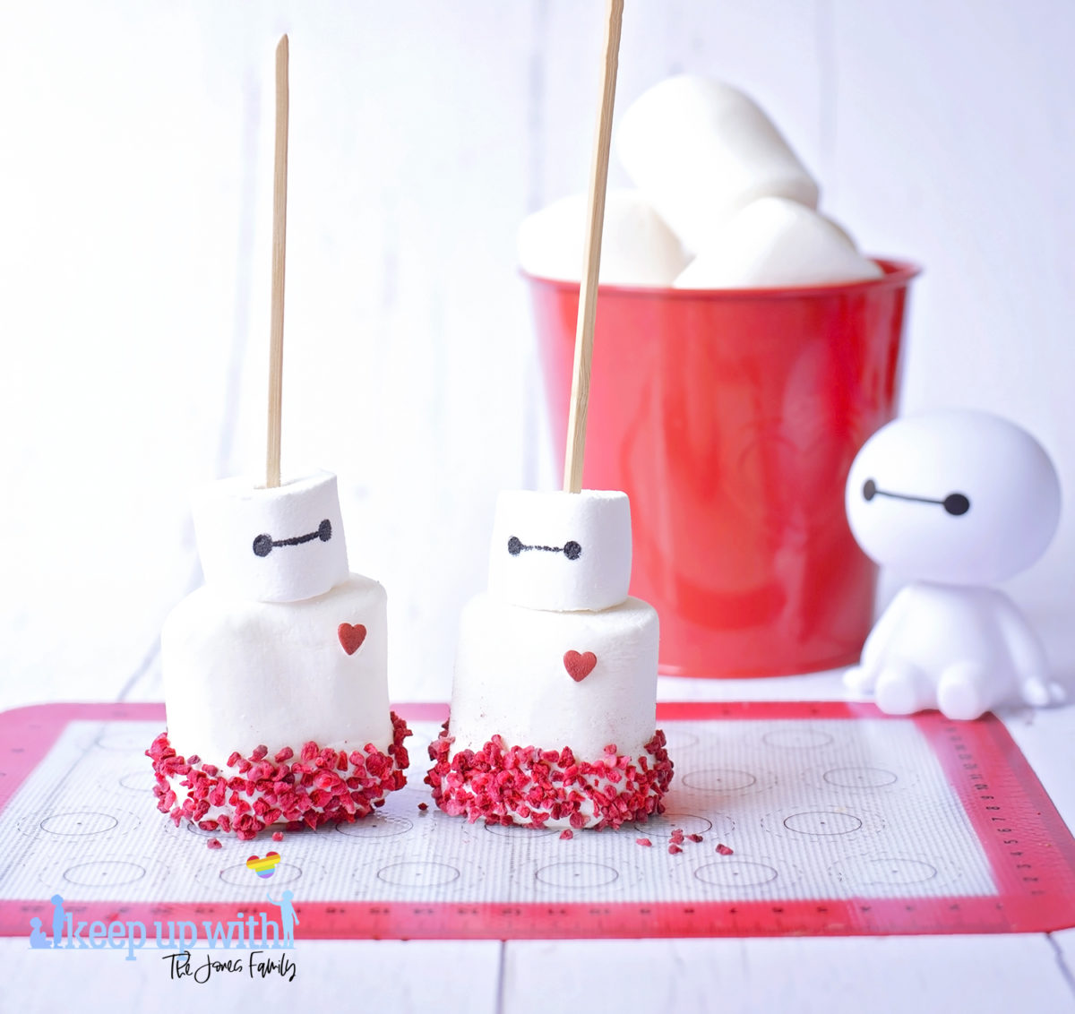 Image shows a plastic Baymax figure from Disney Pixar's Big Hero 6 sat on a white tabletop. In front of him are two fruity baymas marshmallow pops sat on a silicone baking mat. In the background is a red tin bucket of large marshmallows. Image by Keep Up With The Jones Family.