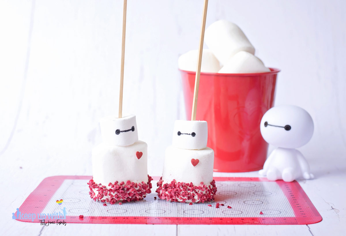 Image shows a plastic Baymax figure from Disney Pixar's Big Hero 6 sat on a white tabletop. In front of him are two fruity baymas marshmallow pops sat on a silicone baking mat. In the background is a red tin bucket of large marshmallows. Image by Keep Up With The Jones Family.