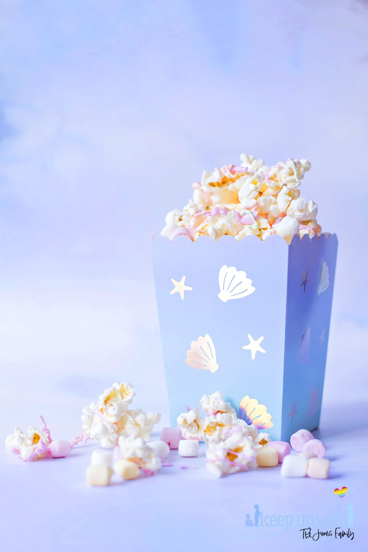 Image shows a carton of Mermaid Popcorn on a pale blue background.  The popcorn is coated in pastel pink, orange and green candy melts, and there are mini pastel marshmallows and pearl sprinkles mixed in.  The popcorn box is mermaid themed from Meri Meri. Image by Sara-Jayne from Keep Up With The Jones Family.