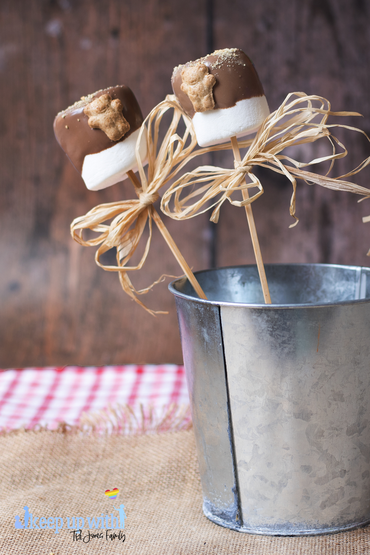 Image shows two teddy bear s'more pops with raffia ribbons in a tin bucket on a dark wooden tabletop. Image by keep up with the jones family.